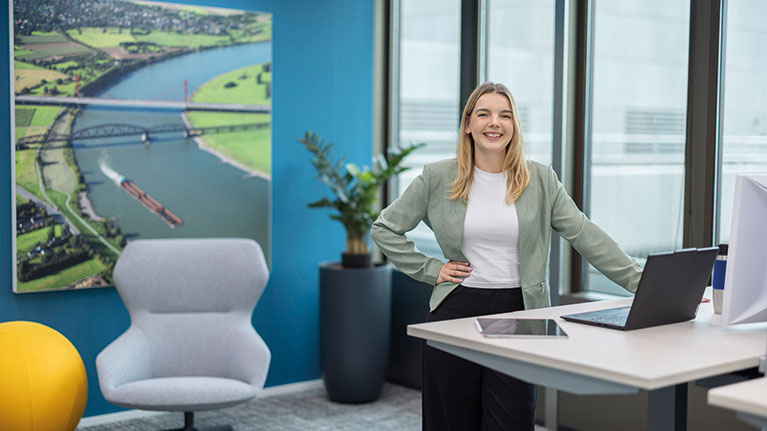 Annika steht an einem Hubschreibtisch mit Laptop vor einem Bild des Rheins.