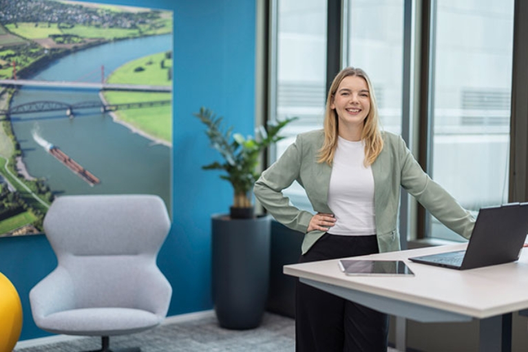 Annika steht an einem Hubschreibtisch mit Laptop vor einem Bild des Rheins.
