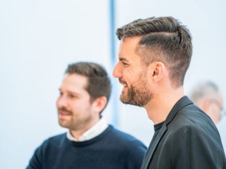 Zwei Männer im Gespräch, einer im Vordergrund im Profil, beide lächeln.