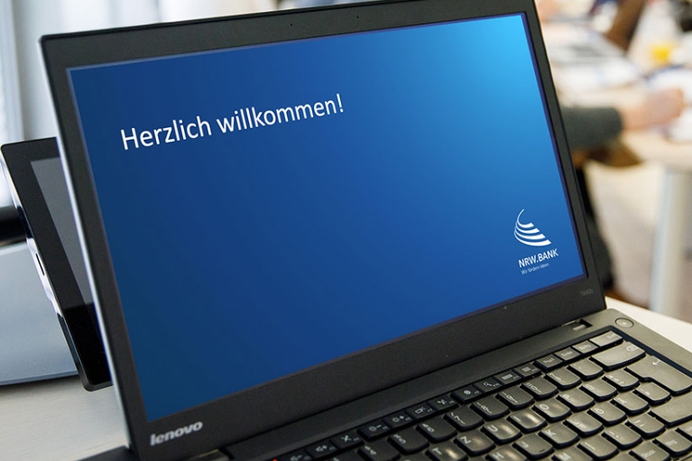 Auf dem Bildschirm eines Laptops steht mit weißer Schrift auf blauem Hintergrund: "Herzlich willkommen!"