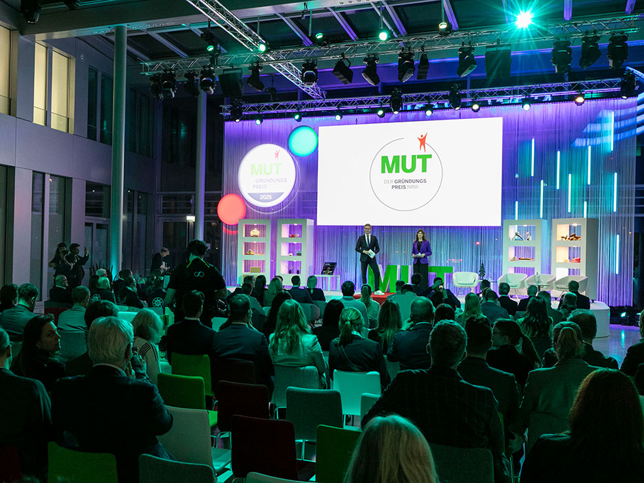 Ein Publikum blickt auf die Bühne, wo zwei Moderierende unter Lichtinstallationen MUT - DER GRÜNDUNGSPREIS präsentieren.