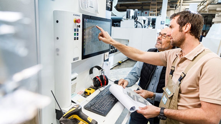 Zwei Männer in einer Werkshalle vor einer modernen Maschine. Einer deutet auf den Bildschirm der Maschine.
