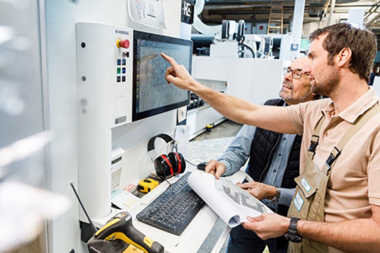 Zwei Männer in einer Werkshalle vor einer modernen Maschine. Einer deutet auf den Bildschirm der Maschine.