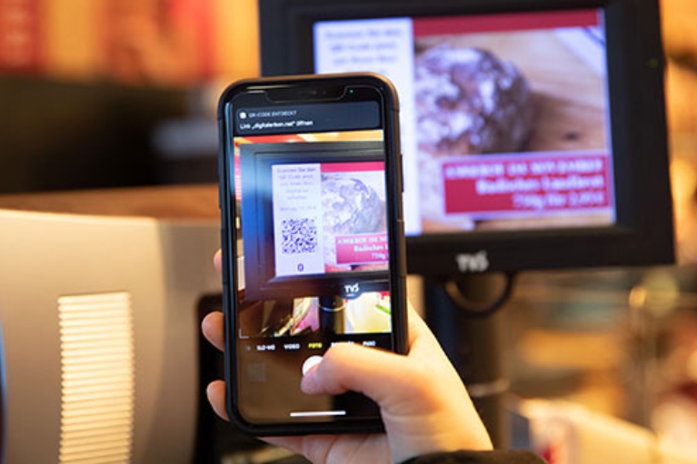 Bildschirm einer modernen Kasse, davor ein Smartphone, das den QR-Code auf dem Bildschirm scannt.