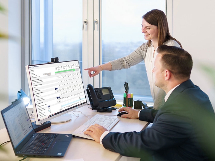 Eine Frau und ein Mann an einem Schreibtisch im Büro. Die Frau deutet mit dem Finger auf eine Tabelle auf dem Monitor.