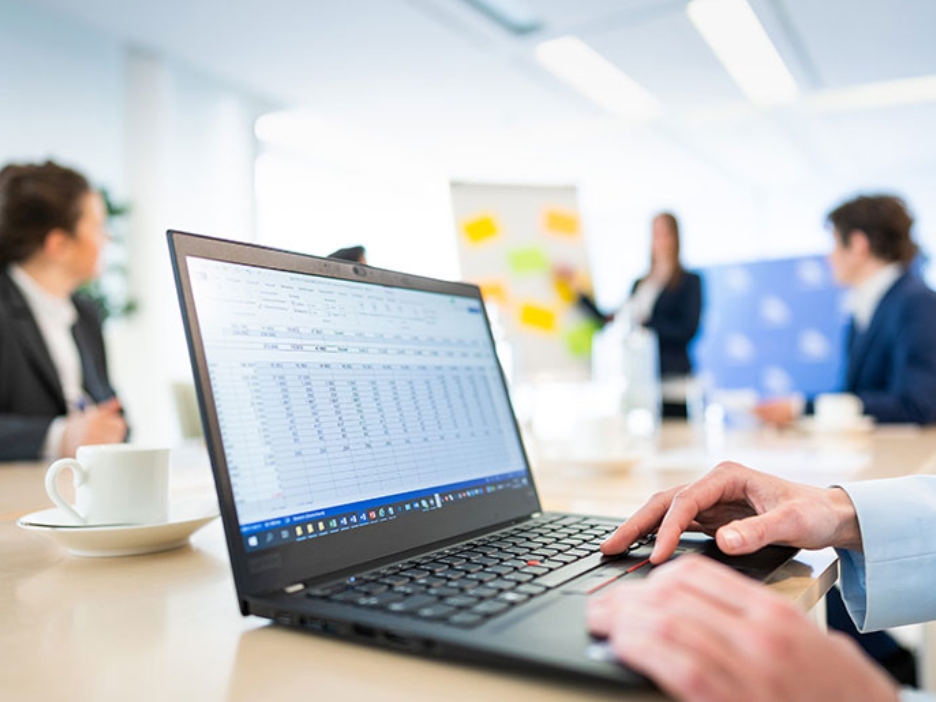 Vier Personen bei einer Präsentation in einem Besprechungsraum. Im Vordergrund ein Laptop mit einer Tabelle