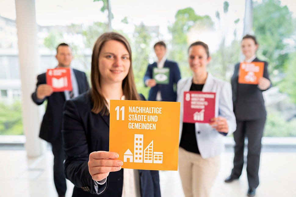erschiedene Mitarbeiter der NRW.BANK halten Karten mit Symbolen der Sustainable Development Goals hoch. Die meisten sind unscharf, auf dem vordersten, welches von einer Frau hochgehalten wird, steht jedoch: „Nachhaltige Städte und Gemeinden“.