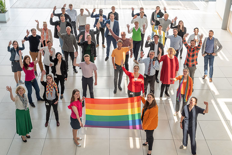 Bunt gekleidete Mitarbeiterinnen und Mitarbeiter der NRW.BANK halten einen Regenbogen-Fahne hoch