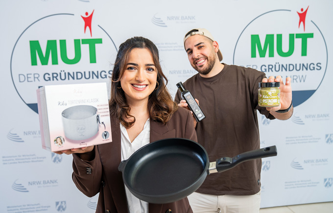 Kiki Aweimer und Hamza Aweimer von Kikis Kitchen vor dem MUT - DER GRÜNDUNGSPREIS NRW-Logo