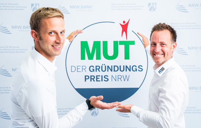 Christian Plett und Florian Eickelmann von PETec Table Lift Systems vor dem MUT - DER GRÜNDUNGSPREIS NRW-Logo