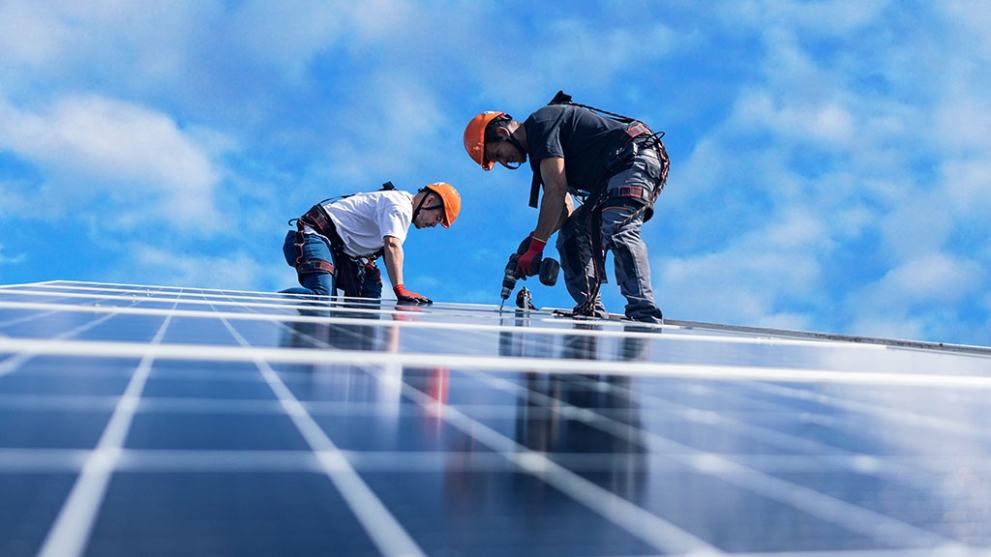 Zwei Männer mit orangenen Helmen befestigen mit einer Bohrmaschine Solarpaneele auf einem Dach´.
