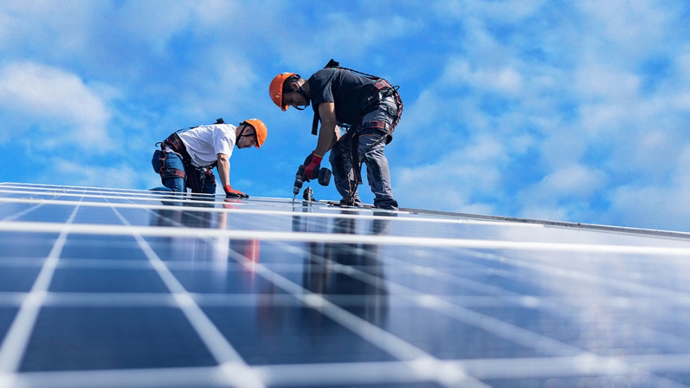 Zwei Männer mit orangenen Helmen befestigen mit einer Bohrmaschine Solarpaneele auf einem Dach´.