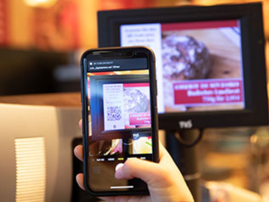 Display einer modernen Kasse, davor ein Smartphone, welches den Code auf dem Display scannt