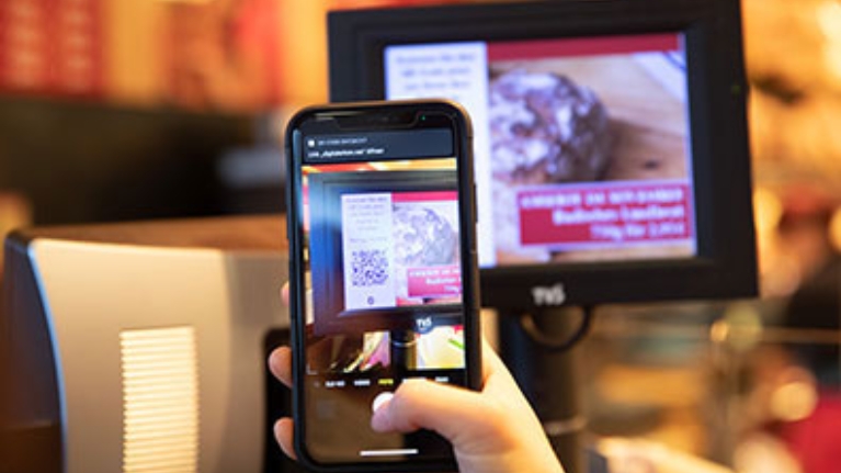 Display einer modernen Kasse, davor ein Smartphone, welches den Code auf dem Display scannt