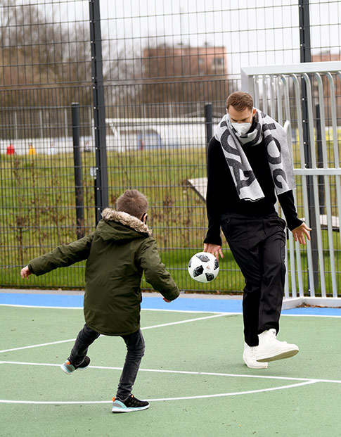 Ein Junge spielt mit Manuel Neuer auf einem Kunstrasenplatz Fußball.