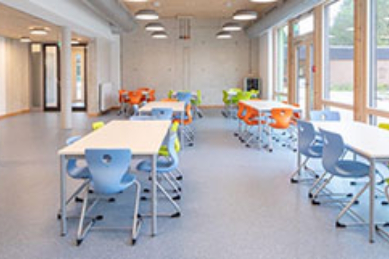 Blick in einen hellen Aufenthaltsraum in einer Schule mit bunten Stühlen und weißen Tischen.