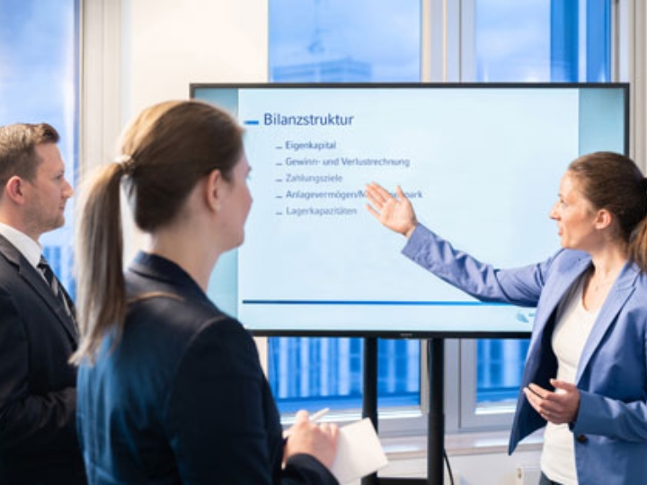 Auf einem Monitor steht das Wort Bilanzstruktur. Eine Frau erklärt etwas dazu.