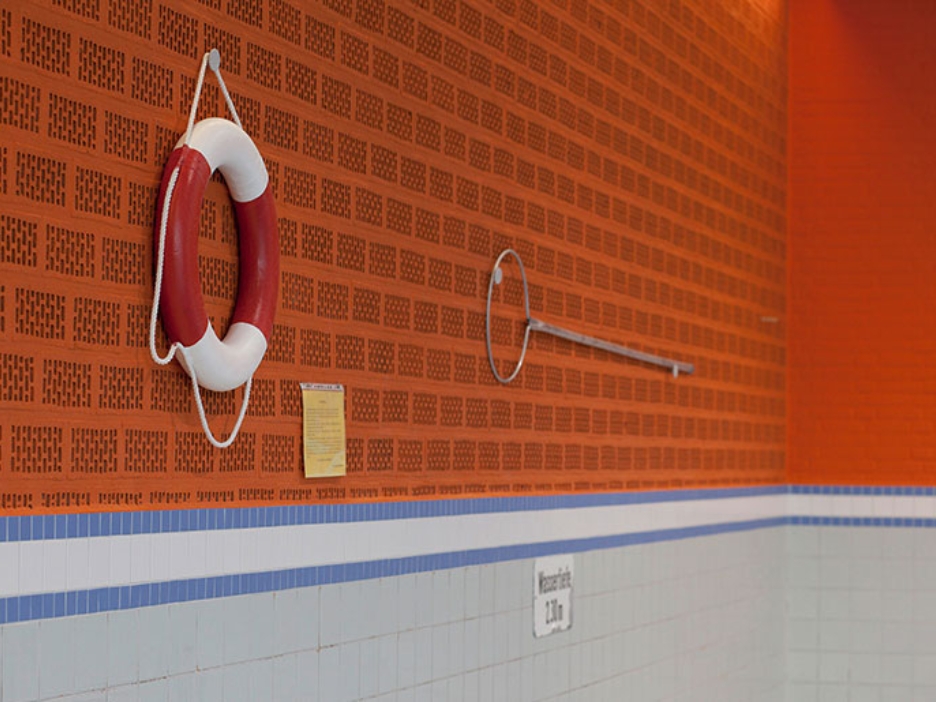 Orangefarbene Wand eines leeren Schwimmbads. An der Wand hängt ein Rettungsring und eine Rettungsstange