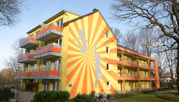 Orange-gelbe Fassade eines Studentenwohnheims mit aufgeklebten Silhouetten von Sportlern
