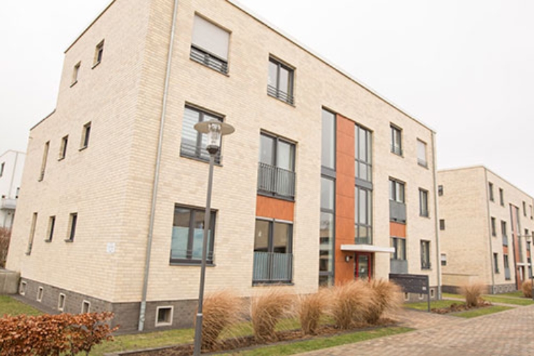 Zwei Gebäude eines Wohnquartiers mit heller Fassade, davor eine ruhige Straße und Vorgärten