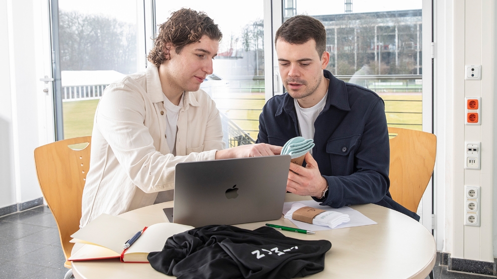 Zwei Männer sitzen am Tisch mit Laptop und Mustern, betrachten gemeinsam ein Paar Socken.