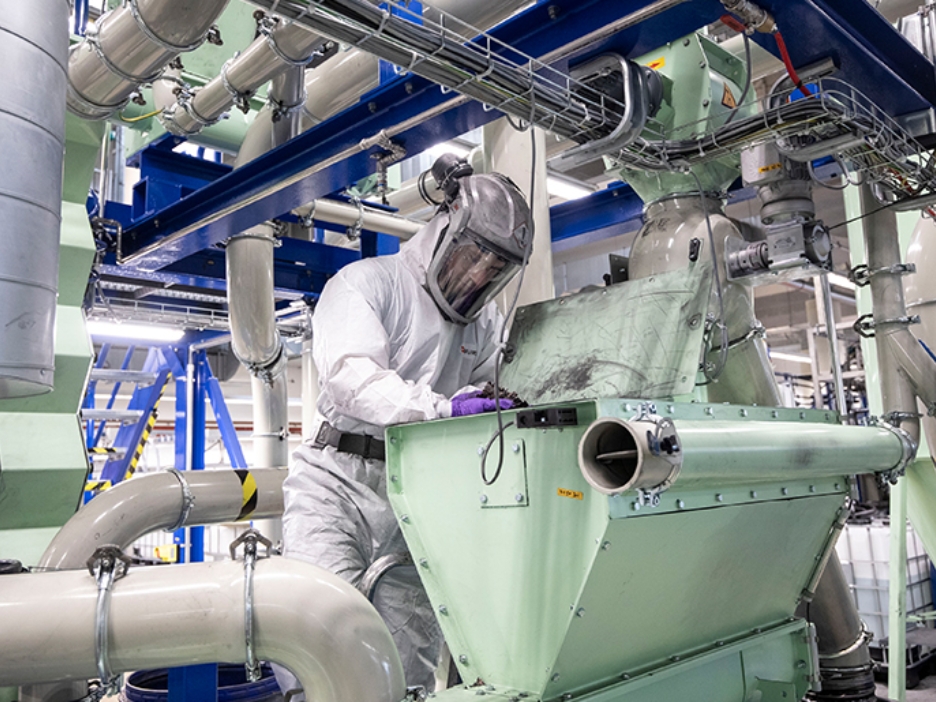 Eine Person in Schutzanzug und Helm arbeitet an einer großen Industrieanlage mit Rohren und Maschinen, beugt sich über einen geöffneten Behälter.