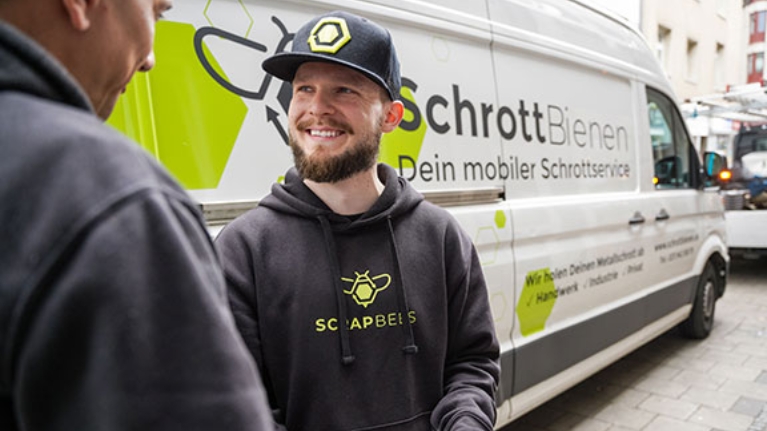 Mann im Scrapbees-Hoodie und Cap lächelt im Gespräch, im Hintergrund ein Transporter mit Firmenlogo.